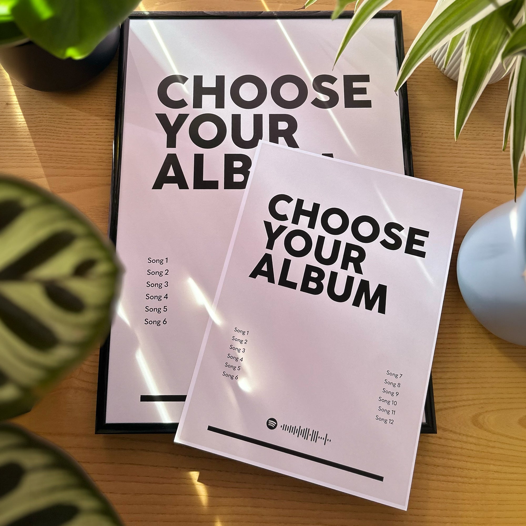 Two customizable album cover mockups displayed on a wooden table with decorative plants. The larger poster in the background reads 'CHOOSE YOUR ALBUM' with a song list, and the smaller mockup in the foreground shows the same design with a white-to-pale-pink gradient.