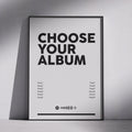 A framed minimalist poster leaning against a light gray wall on a floor, with large black text reading 'CHOOSE YOUR ALBUM' and small song titles arranged in two columns below.