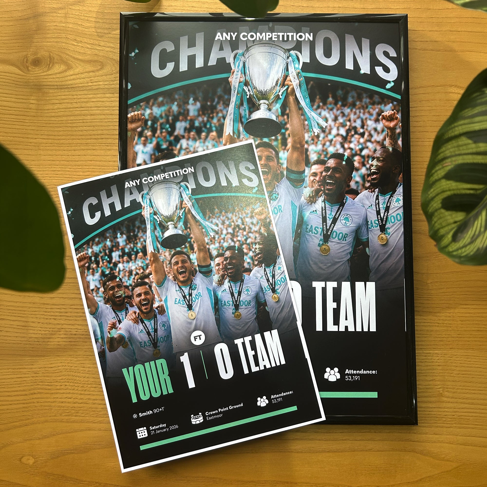 Two customizable sports championship posters displayed on a light wood tabletop, with green leaves framing the sides. The larger poster in the background features a team celebrating with a trophy, and the smaller poster in the foreground shows a similar design with placeholder text.