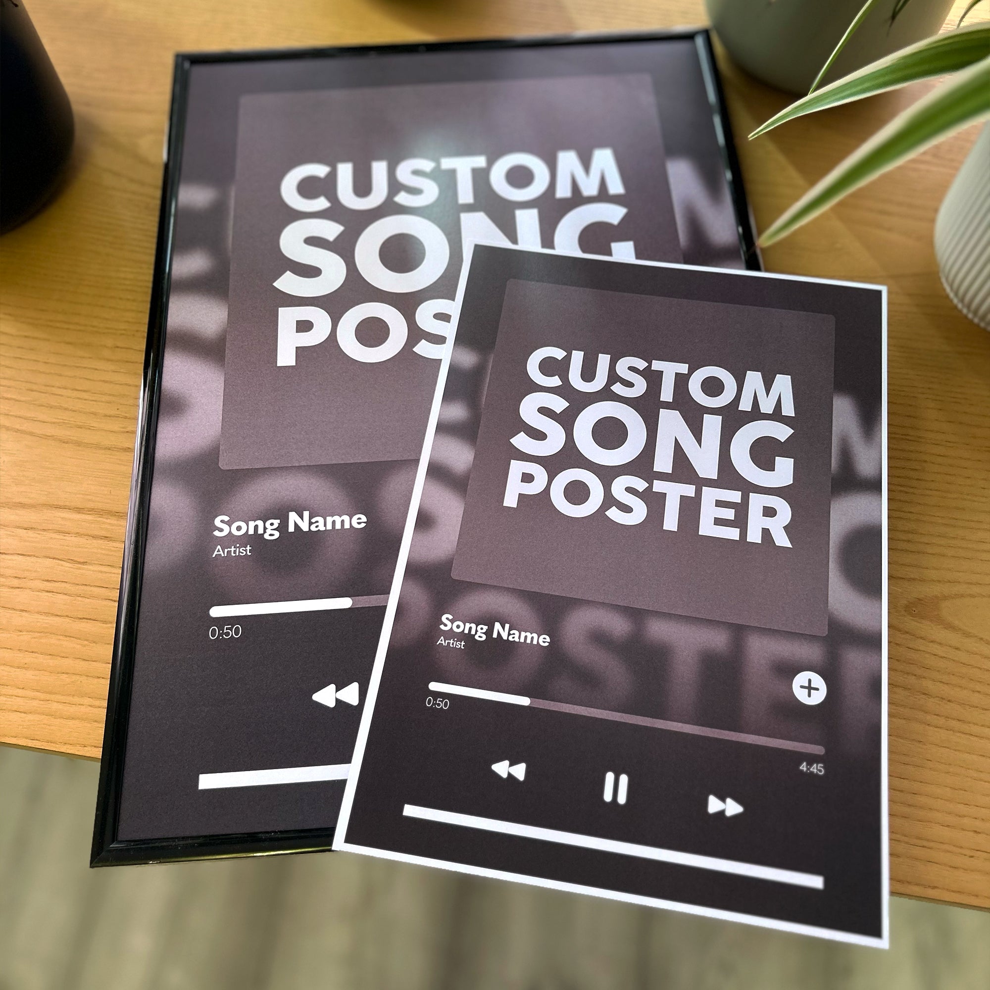 Two custom song poster prints displayed on a wooden tabletop, one in a black frame and one flat, both featuring a dark music player interface with white text placeholders.