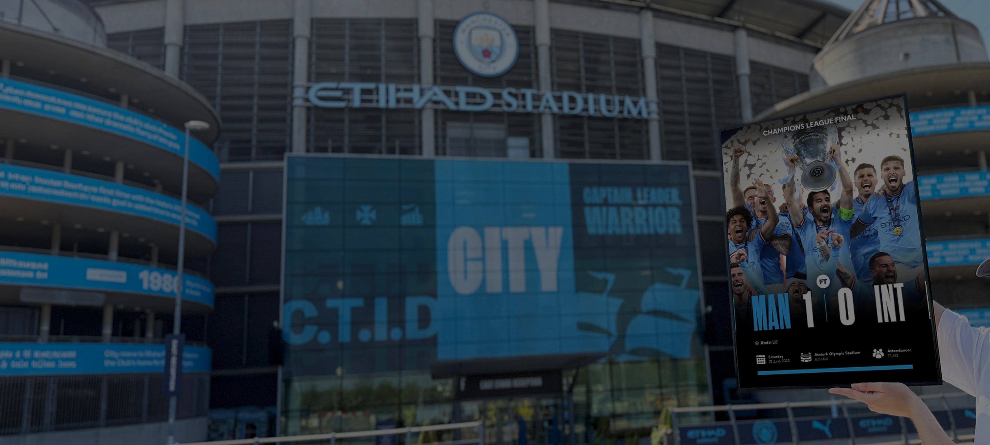A large digital display at Etihad Stadium shows the text 'CITY' with a football player in a blue jersey, while a hand holds a tablet displaying a match score of 'MAN 1-0 INT'.